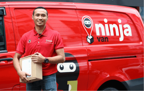 Delivery person with Ninja Van branded red uniform standing next to delivery van holding a package, promoting logistics and courier services.